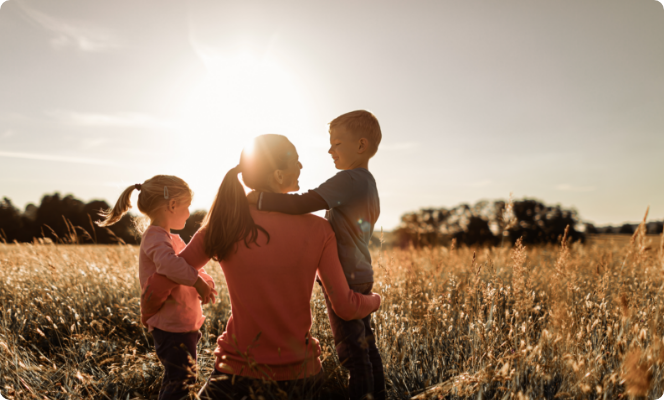A mother kneeling in a field during sunset, holding her young son on one side and her daughter on the other, enjoying family time outdoors.