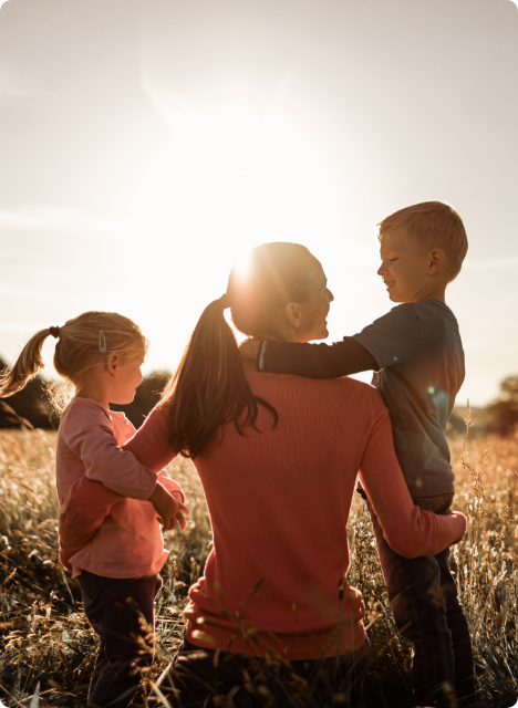 A mother kneeling in a field during sunset, holding her young son on one side and her daughter on the other, enjoying family time outdoors.
